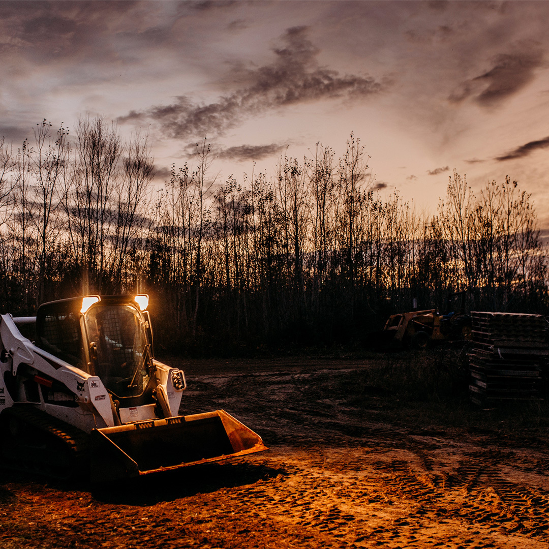 A skid steer sits in a cleared field. Tread tracks mark up the ground below a skid steer. The skid steer has a low profile bucket attached. The sun sets in the background